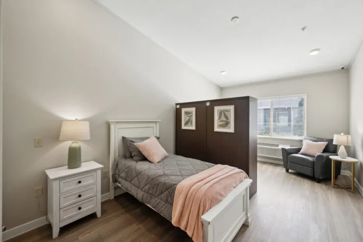 Private bedroom with soft lighting, comfortable bed, seating area, and natural window light., Harvard Square Memory Care