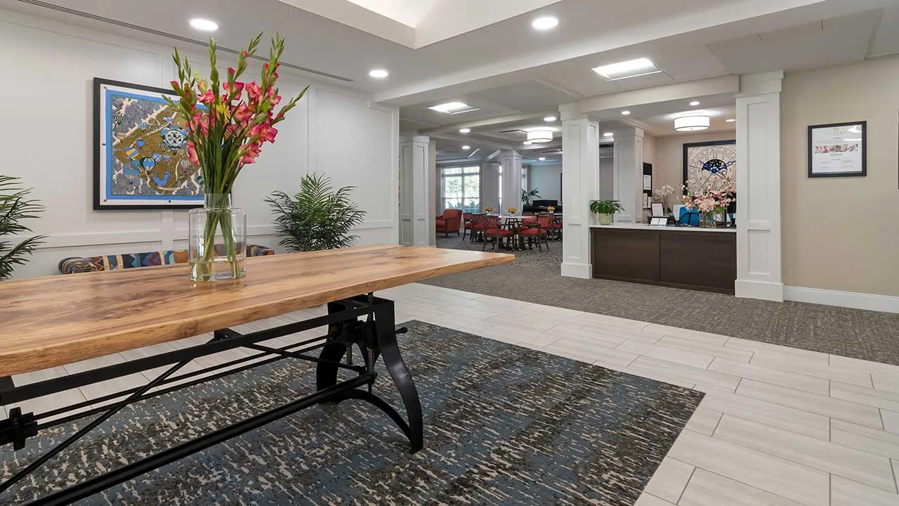 A brightly lit, modern lobby with a wooden table topped with a vase of flowers in the foreground. There are plants and framed artwork on the walls. In the background, there's a reception desk and a seating area with red and brown chairs.