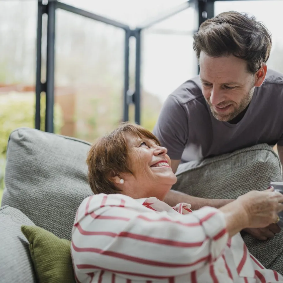 A smiling elderly woman receives a warm drink from a caring man in a bright living room.