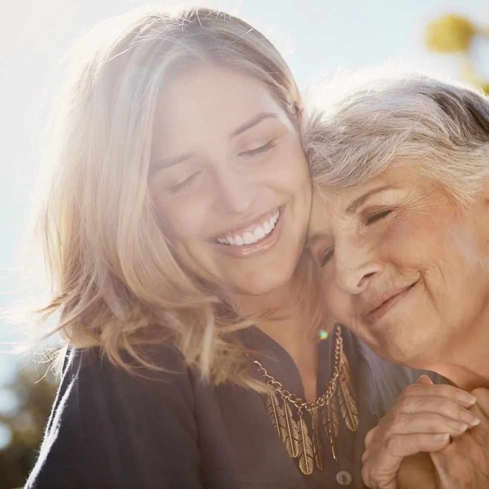 A senior woman and younger woman share a warm, joyful embrace outdoors in sunlight.