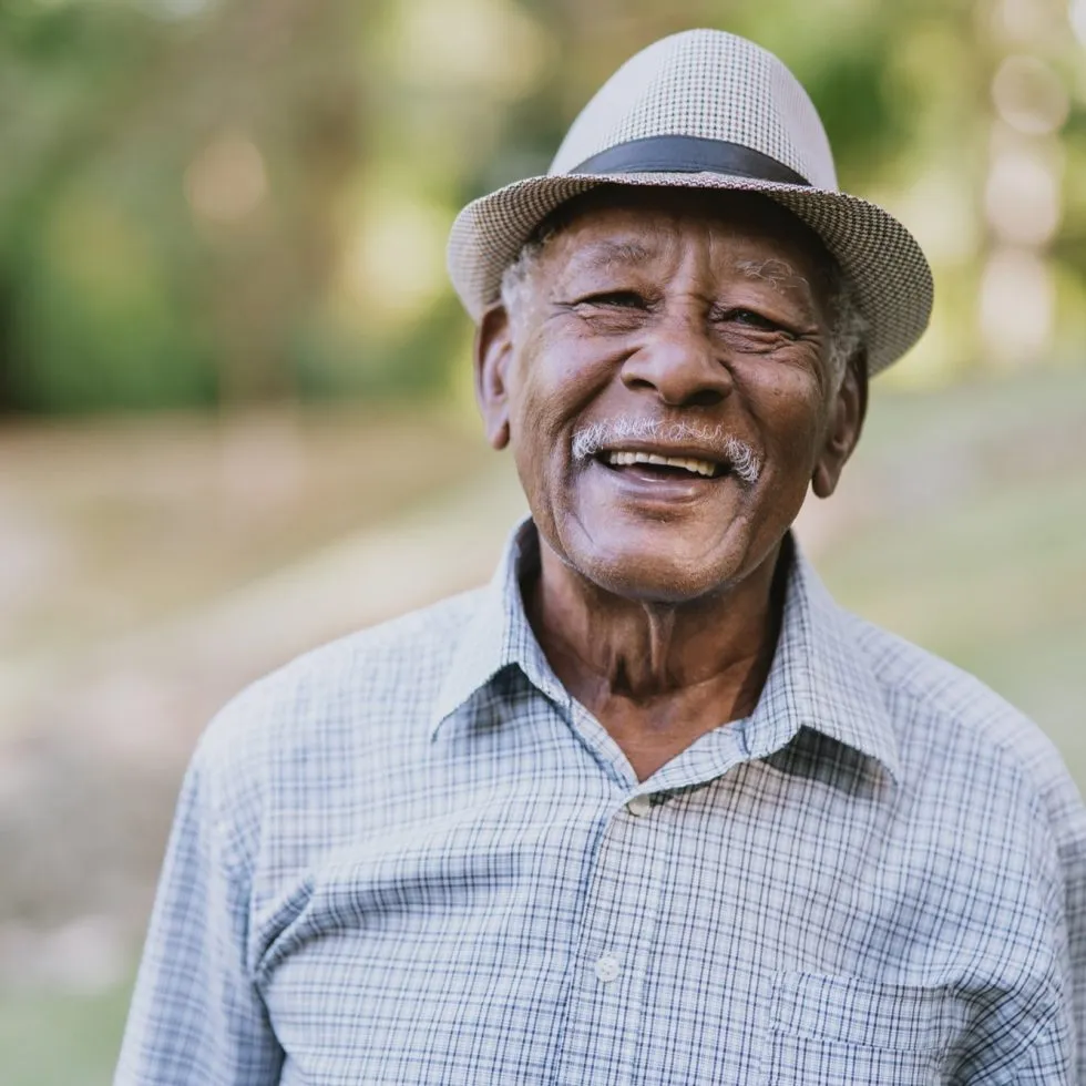 Smiling elderly man wearing a hat enjoying a peaceful outdoor setting in nature.